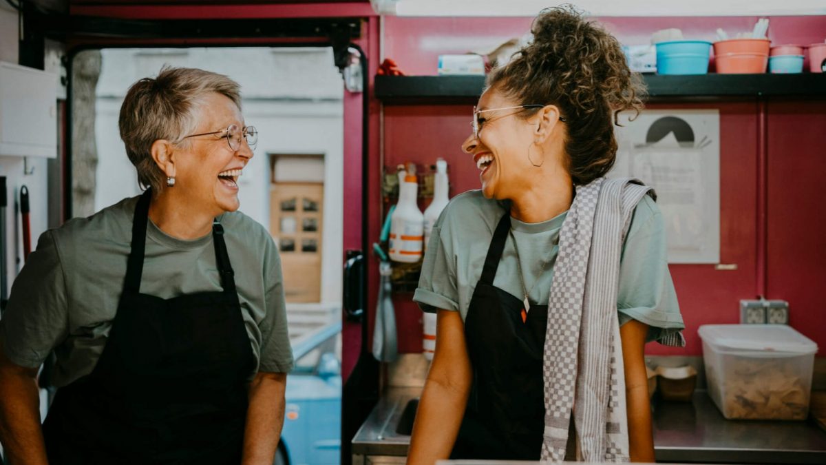 Duas mulheres empreendedoras rindo apoiadas na bancada de atendimento de uma lanchonete - negócio e empreendedorismo